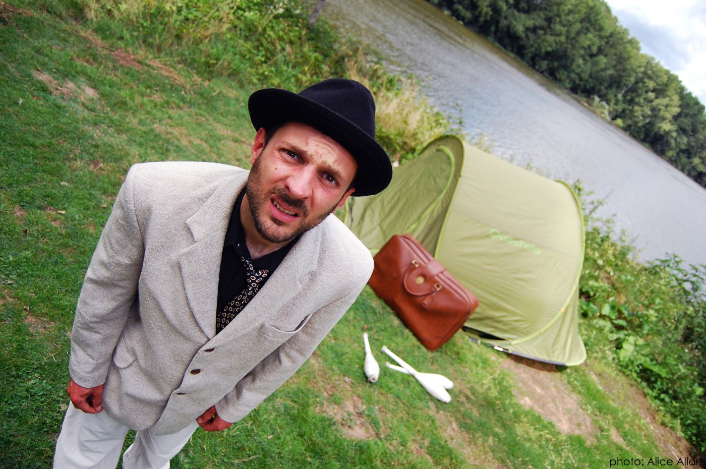 Artiste vêtu d’une veste trop grande qui regarde l’objectif. En arrière-plan, une tente de camping avec une valise et des massues à l’entrée, ainsi qu’une rivière.