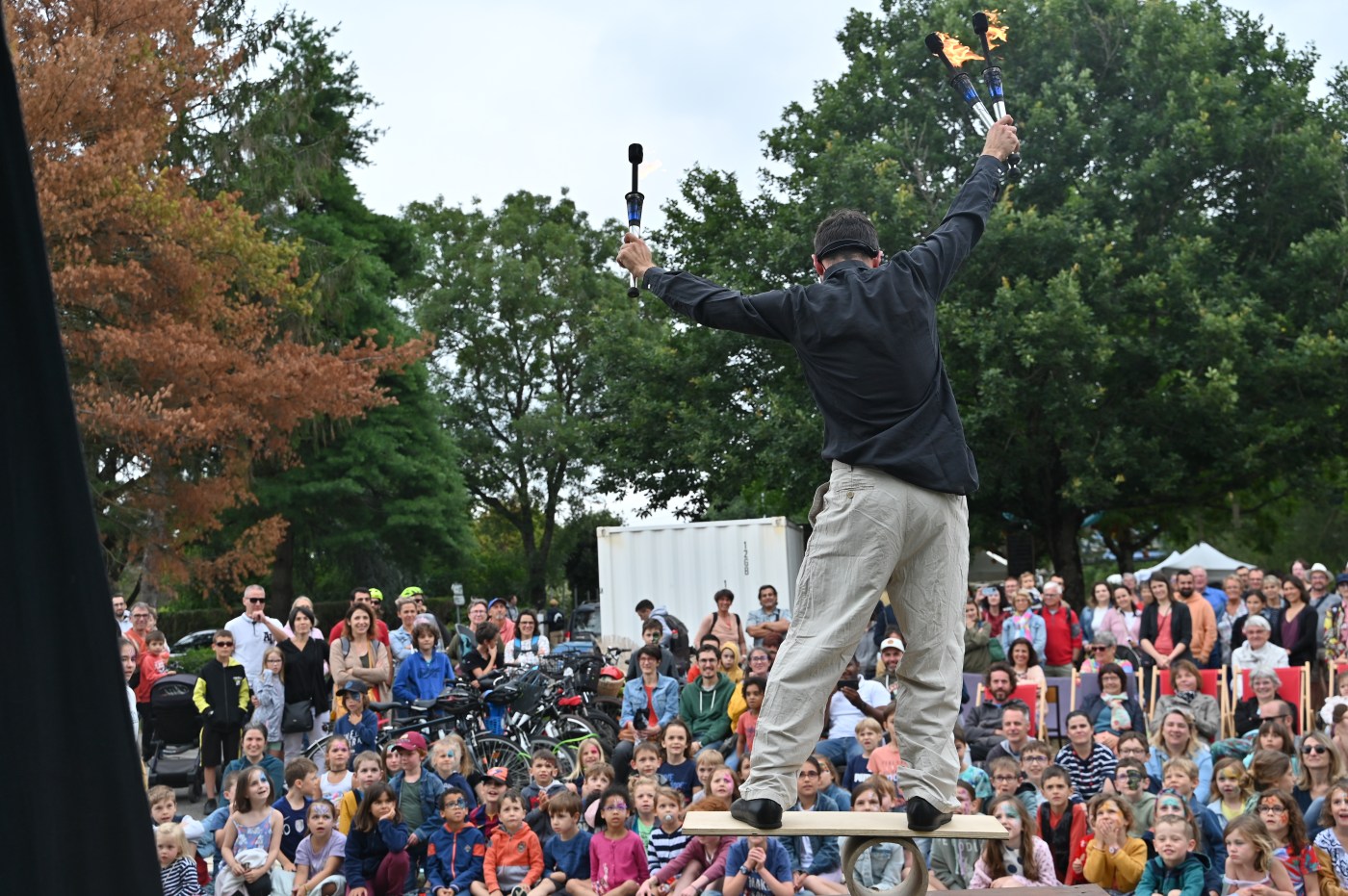 Artiste de cirque sur un rola-bola, jonglant avec des torches enflammées, face à un public amusé et émerveillé lors d’un spectacle en extérieur.